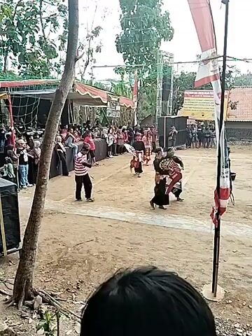This is a medium-shot video taken outdoors, likely in a village or rural setting, during daylight. The scene captures what appears to be a cultural performance or festival. In the foreground, a blurry out-of-focus human head is visible at the bottom of the frame. The main area of action is a dusty, open ground with a white line dividing it.

On the left side of the frame, a large tree trunk with exposed roots is prominently featured, partially obscuring the background. Behind the tree, a section of a stage or performance area is visible, covered by a red and yellow awning and with netting stretched above. A crowd of spectators is seated and standing behind a low wall or barrier in this area.

In the center of the open ground, several individuals are engaged in a performance. One person, wearing a striped shirt and dark pants, is holding a whip-like object. Two other performers, dressed in dark clothing and wearing elaborate headdresses or costumes that resemble animal heads (possibly horses or mythical creatures), are also active. Another individual in dark attire is walking across the scene. Further to the right, a person in dark clothing with a white and red sash is standing. In the background, more people are gathered, and there are structures that suggest tents or temporary buildings.

To the right of the performance area, a tall pole with red and white flags is erected. Behind this, a large speaker system is visible, along with more tents and buildings, including a tiled roof structure. The overall impression is of a community event taking place in an informal, outdoor venue. The atmosphere seems lively, with performers actively engaged in their activity and an audience observing.