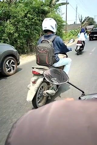 The video shows a first-person view from a motorcycle traveling on a paved road. In front, another motorcycle is being ridden by a person wearing a white helmet and a backpack. The rider in front is wearing a blue shirt and light-colored pants. There are houses and trees visible on the left side of the road, and utility poles with wires stretch overhead. Further ahead, a black pickup truck and another motorcycle can be seen on the road. The camera is pointed forward, capturing the scene from the perspective of the second motorcycle. The overall environment appears to be a rural or suburban area with daylight.