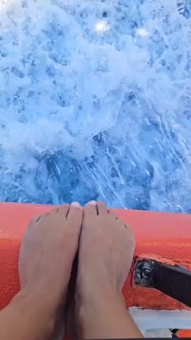 A pair of bare feet are visible, resting on the orange edge of a boat. The boat is moving through blue water, creating white foam and splashes as it goes. The camera is looking down at the feet and the water. In the distance, land with hills is visible.