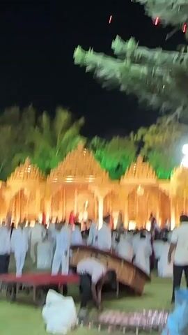 The video shows an outdoor nighttime gathering, likely a wedding reception. In the background, a large, ornately decorated stage-like structure is illuminated with warm-toned lights and green spotlights, creating a visually striking focal point. A crowd of people, mostly men, dressed in white shirts and pants, are gathered around the structure, possibly watching or participating in an event. In the foreground, a man is bending over what appears to be a large, decorative tray or platform, possibly arranging or cleaning up. A small, red cart is nearby. Above, red fireworks or similar pyrotechnics are visible in the dark sky, adding to the festive atmosphere. A large tree branches into the shot from the top, partially obscuring the view.