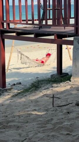 A person wearing a red hat and red swim trunks lies in a hammock on a sandy beach. The hammock is suspended between two bamboo poles. Behind the hammock, a wooden structure is visible, as are flags on poles. The ocean and a distant cityscape can be seen in the background. The foreground is dominated by sand and some sparse vegetation. The lighting suggests it is daytime.
