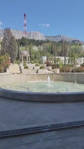 The scene captures a well-maintained outdoor space featuring a multi-tiered fountain as its central element. The fountain is a circular structure with several smaller fountains spraying water upward, creating ripples on the water surface. Behind the fountain, there's a landscaped area featuring a gazebo, steps, plants, and neatly trimmed hedges, giving the space a garden-like aesthetic. In the background, a mountain range dominates the horizon, with a tall, striped communications tower prominently positioned on the peak. The sky is clear and blue, indicating sunny weather. The ground surrounding the fountain is paved, and there's a drainage system visible along the edge. Overall, the scene conveys a sense of tranquility and constructed beauty, possibly within a park or garden setting. There are paint buckets in the corner of the scene.