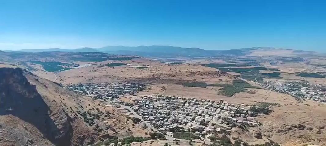 This is a drone footage video taken in the Galil Tachton Regional Council, Israel. The footage shows a panoramic view of a mountainous and dry landscape under a clear blue sky, suggesting a sunny day with good weather. The scene features rugged, eroded cliffs and hills, typical of arid or semi-arid regions. In the mid-ground, a small town or village is visible, nestled amongst the hills. The buildings appear to be densely packed, with light-colored roofs. The overall impression is one of a remote, natural environment with human settlement integrated into the landscape. The camera moves and pans, offering a dynamic perspective of the terrain, highlighting the vastness and rugged beauty of the area. There are no people or specific objects clearly discernible in the footage, the focus is on the landscape and the settlement. The time of day appears to be daytime, as indicated by the bright sunlight and clear visibility. The location cues, such as the arid landscape and mountainous terrain, are consistent with descriptions of regions within Israel. The video does not contain any visible text.