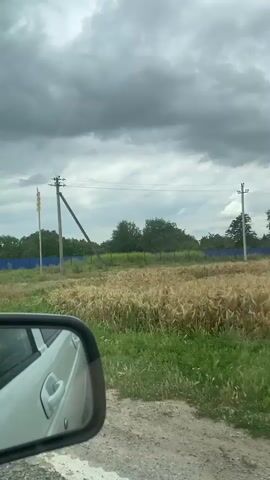 The video appears to be taken from inside a vehicle, likely a car, as evidenced by the reflection in the side-view mirror. The perspective suggests the vehicle is moving along a dirt or gravel road, with a grassy area and some vegetation visible to the right. In the distance, there are trees and a blue fence. A traffic light is visible on a pole, and a tractor with a trailer is passing by. The sky is overcast with dark clouds, suggesting the possibility of rain. The overall environment seems rural or semi-rural.