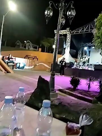 A nighttime outdoor scene features a band performing on a stage illuminated by purple lights. A singer stands center stage, while other band members play instruments. In the foreground, a decorative lamppost stands next to a tree. Several plastic water bottles are visible on the ground in the lower left corner. The background shows a terraced seating area and building structures, suggesting a public gathering space or park.