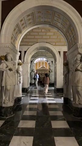 The video shows a long, marble-tiled corridor with arches and checkered black and white flooring. Several people are walking through the corridor towards a central altar-like structure at the far end. On either side of the corridor, there are large marble statues. The ceiling of the corridor features a repeating pattern of gold stars on a yellow background. The lighting is dim, creating a solemn and majestic atmosphere.