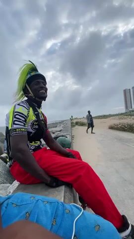 A person with bright green hair styled into a mohawk under a helmet sits on a concrete barrier, looking to their right and smiling. They are wearing a black and neon yellow cycling jersey with various logos and bright red pants. Rollerblades are attached to their feet. In the background, a person walks away from the camera on a paved path near a sandy beach. In the distance, tall buildings are visible. The sky is cloudy. The lower portion of another person, wearing blue pants with a bee pattern, is visible in the foreground.