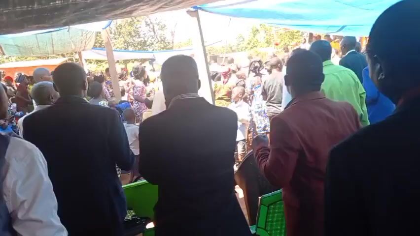 A crowd of people is gathered under a blue and red tarp structure outdoors. Most of the people in the foreground have their backs to the camera, and they appear to be watching something happening at the front of the crowd. Many are wearing suits or other formal attire. A few children are visible in the crowd. The sky is bright and the sun is shining.