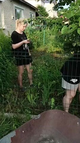 A young woman with blonde hair pulled back in a ponytail, wearing a black t-shirt and white shorts, stands in a yard near a brick building. She is holding a smartphone and looking down at it. A wire fence separates her from the foreground, where part of a rusty wheelbarrow is visible. Behind her, the yard is overgrown with tall green grass and bushes. Sunlight illuminates the scene. The woman then bends down, reaching towards something on the ground near the fence.