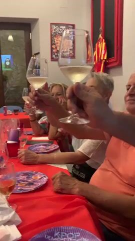 A group of people are seated around a red-covered table, raising glasses filled with a light-colored liquid in a toast. The individuals are of various ages and genders, some are smiling, and one child is visible. The room appears to be a pizzeria, with pizza-themed decorations on the wall, including a large mural of pizzas. Red plastic cups and paper plates with colorful designs are also on the table. A man in a white shirt is standing and pouring liquid into glasses. The lighting is warm and inviting.