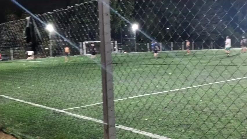 A group of men are playing soccer on a green field at night under bright spotlights. The game is viewed through a chain-link fence. A person in an orange and black soccer jersey walks past the fence and looks down. Other players are visible on the field.