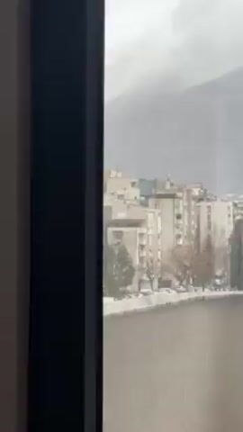 A medium shot through a window of a European cityscape on an overcast day. Several concrete apartment buildings are visible, as are trees without leaves and some cars in a parking lot. Behind the city, there is a large mountain shrouded in fog. Snow is falling lightly. The window frame is black.