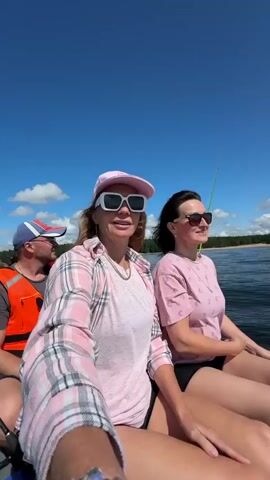 A video shows three people on a boat under a bright blue sky with scattered clouds. In the foreground, a woman wearing a pink plaid shirt, a pink baseball cap, and white sunglasses smiles at the camera. To her right, another woman in a pink t-shirt and sunglasses looks off to the side. Partially visible behind them on the left is a man wearing an orange life vest and a blue baseball cap. The boat is moving through dark water, leaving a wake. A treeline is visible in the distance across the water.