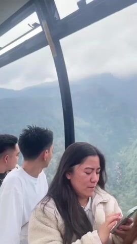 The video shows a group of people inside a cable car with a scenic view of mountains covered in mist and trees. In the foreground, a woman with long dark hair is looking down at her smartphone. She is wearing a cream-colored jacket over a white shirt. To her left, a man with short dark hair is looking out the window, and next to him, another man in a white t-shirt is also looking away from the camera. The cable car's interior is visible, with part of the window frame and a yellow safety strap. The background reveals layers of green mountains shrouded in a misty atmosphere, with a small river or stream visible in the valley below. The overall impression is of a journey through a lush, mountainous landscape.