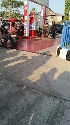 A group of people on motorcycles are waiting in line at a gas station. The gas station has red and white branding and several gas pumps. The sun is shining, casting shadows on the ground. Some people are wearing helmets.