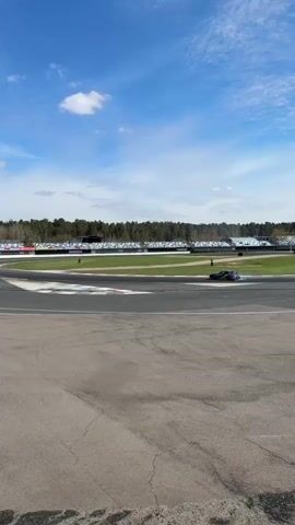 The video shows a purple, green, and black car drifting on a racetrack, creating a cloud of smoke. The track is surrounded by grass, tires, and a barrier. In the background, there is a grandstand, a line of trees, a light tower, and a blue sky with light clouds. There are also people in the background. The camera is still and captures the car drifting around a turn in the track.