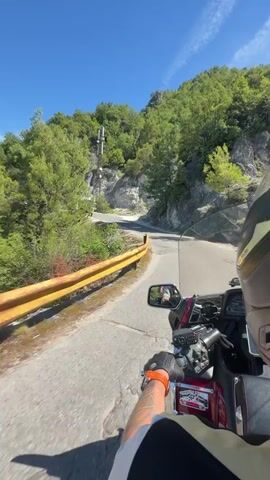 A first-person perspective video shows someone riding a motorcycle on a paved road. The motorcycle is red and black. The rider is wearing a black helmet, black gloves with orange accents, and a black and white jacket. The road curves to the left and ascends a hill. To the left of the road is a yellow guardrail and a steep, rocky incline covered with green trees and bushes. To the right of the road is a similar incline covered with trees and bushes. The sky is clear and blue. In the distance, a telecommunication tower is visible on the left side of the road, near a tunnel entrance. Another motorcycle is visible further up the road, approaching the tunnel.