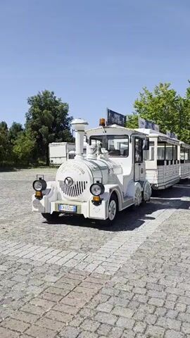 A white toy train sits on a cobblestone surface under a clear blue sky. The train has a locomotive at the front and several passenger cars behind it. The locomotive is white with a black grill and two headlights. It also has a tall smokestack and a small roof with orange lights. The passenger cars are also white and have windows. The train is parked in an outdoor area with trees and buildings in the background. The video is taken from a low angle, looking up at the train.