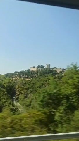 A blurry video captures a distant stone fortress perched atop a lush green hill under a clear blue sky. The camera appears to be moving, likely from a vehicle, as the trees in the foreground are blurred with motion. The fortress itself is built of light-colored stone and features several towers and a long wall. It is surrounded by dense vegetation, including trees. The overall impression is of a historical site situated in a natural landscape, viewed from a distance.