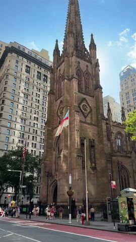 The video shows a stone church with a tall steeple against a bright blue sky with some clouds. A flag with red and white stripes and a blue canton is flying on a flagpole in front of the church. Several people are walking on the sidewalk in front of the church. There are tall buildings in the background. The sun is shining brightly and there is lens flare.