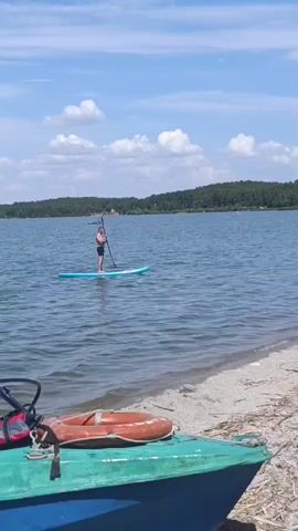 A man stands on a paddleboard in the water and paddles with a paddle. The paddleboard is blue. The man is wearing black shorts. The water is blue and has waves. The sky is blue with white clouds. There are trees in the background. A beach with sand and some dried vegetation is in the foreground, along with parts of a green and blue boat with a life preserver.
