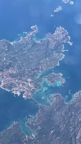 This aerial video presents a stunning daytime view of a rugged coastal landscape, consistent with the description of Porto Cervo, Italy. The scene is dominated by the vast expanse of the deep blue sea, interspersed with brilliant turquoise patches indicating shallow waters and submerged features.

The landmass itself features an irregular, rocky coastline with scattered vegetation, appearing as various shades of green and grey. A significant urban settlement is visible along the coast, characterized by numerous white and light-colored buildings clustered together. A prominent feature within this developed area is a large, busy harbor or port, clearly showing multiple docks and infrastructure, suggesting commercial or recreational marine activity.

Numerous roads can be seen winding through the terrain, connecting different parts of the coast and inland areas. Several small, rocky islands are scattered offshore, some appearing barren while others show traces of vegetation. The sunlight illuminates the scene beautifully, creating clear visibility and highlighting the vibrant colors of the water. The perspective is from a high altitude, likely from an airplane or drone, offering a wide, panoramic view of the intricate coastline and its surrounding waters. No people or discernible text are visible in the video.
