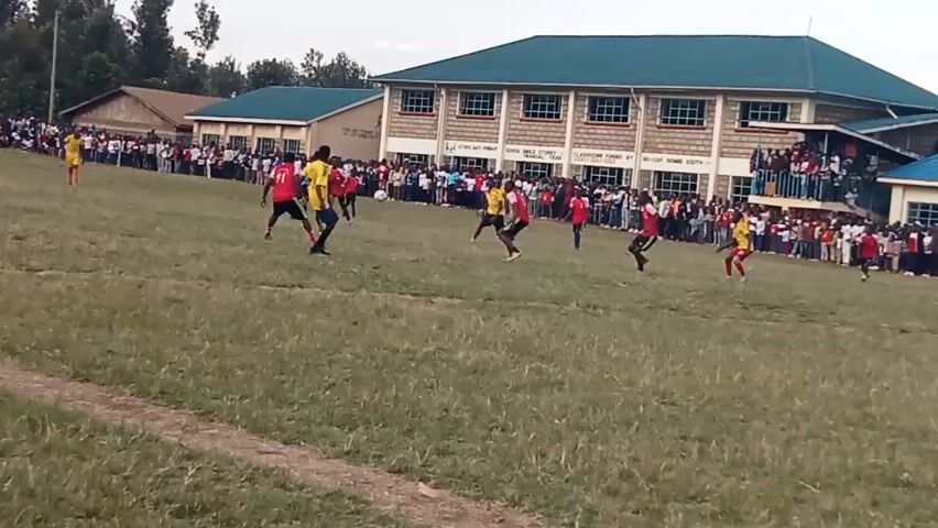 A soccer game is being played on a grassy field. Several players in red and yellow jerseys are on the field, actively engaged in the game. A large crowd of spectators stands behind a barrier, watching the match. In the background, there are several buildings, including a larger, modern-looking building with a blue roof, and smaller structures behind it. The sky appears overcast.