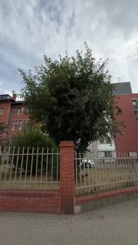 The video opens with a low-angle shot looking up at a brick building with several windows and a light-colored upper section. A metal fence with brick pillars runs across the foreground. A tree with green leaves partially obscures the building. The sky above is cloudy with patches of blue. The scene appears to be an exterior shot of a building in a residential or urban area.