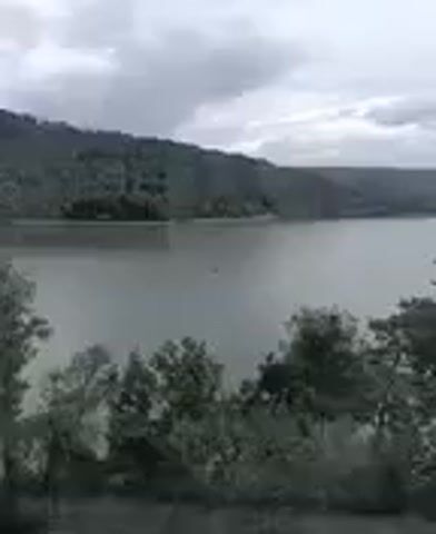This media file shows a wide, calm body of water, likely a lake or a river, with lush, tree-covered hills in the background. The sky is overcast with heavy, gray clouds, suggesting a dim or possibly rainy day. The overall scene is serene and natural, with no visible signs of human activity beyond what might be a small boat far in the distance on the water. The foliage in the foreground is dense and green, with pine trees being prominent. The weather appears cool and possibly damp. The footage seems to be captured from an elevated position, possibly from a building with large windows, as indicated by the dark, vertical frame that appears on the right side of the screen in some frames. There is no visible text within the media file. The setting is identified as Doksy, Czechia, which is known for its natural beauty and lake Máchovo jezero.
