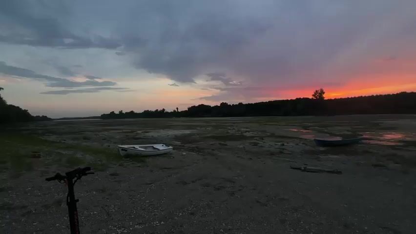 A static, eye-level shot captures a desolate, low-lying area of muddy ground and scattered debris at dusk. A prominent, dark gray electric scooter stands in the lower left corner of the frame. A small, white boat rests on the ground in the middle left, and a dark blue boat is situated in the middle right. The background features a dense line of dark trees silhouetted against a vibrant sky filled with dramatic clouds, tinged with hues of orange, pink, and red from the setting sun. The overall atmosphere is somber and serene, emphasizing the natural beauty of the twilight sky against a stark, neglected landscape.