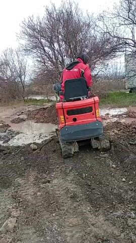 Two men are working in a muddy, rural environment. One man is operating a small, red tracked vehicle. The other man is using a shovel near a large puddle.  A rusted barrel and a hose with a blue object attached are visible nearby. The sky is overcast, and bare trees dot the landscape.