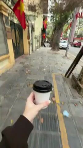 A first-person perspective video shows someone walking along a sidewalk with a cup of coffee. The sidewalk is paved with gray tiles and covered with fallen leaves. A yellow line marks the edge of the sidewalk next to the street. Buildings line the street, some adorned with Vietnamese flags. Cars are visible on the street. The person is wearing a brown sweater and holding a white coffee cup with a black lid.