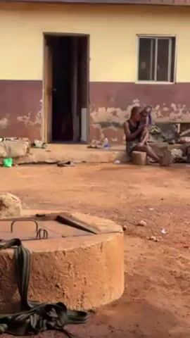 The video shows an outdoor courtyard in front of a building. An adult, possibly female, sits on a small stool holding a young child in their arms. In the background, through an open doorway, a man sits on the floor inside the building. There are sandals outside the door. In the foreground, there is a round, concrete structure that appears to be a well. The ground is reddish dirt. The exterior walls of the building are peeling and partially painted in a light yellow color, with some brown sections.