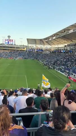 The video captures a scene at a soccer stadium during what appears to be a game or pre-game event. The camera pans across the stadium, showing a large crowd of spectators in the stands, many wearing white and green team colors. A soccer field is visible in the distance, with players on the field and a large video screen displaying images of people in team jerseys. The sky is clear and blue, indicating fair weather. A large digital clock on the side of the stadium shows the time as 18:23. In the foreground, a group of people are walking, some are wearing team apparel, and one person is holding a flag with team colors. The atmosphere appears lively and celebratory.