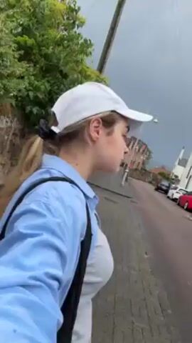 Okay, here is a concise description of the video:

The video shows a young woman walking along a sidewalk on a cloudy day. She is wearing a white baseball cap, a light blue button-down shirt, and a white t-shirt. She has a black bag slung over her shoulder. She is walking on a paved sidewalk beside a street with parked cars. There are houses visible in the background. The video appears to be shot with a phone, moving with the woman as she walks.