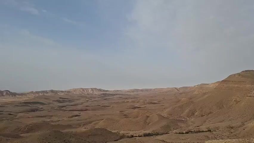 The video depicts a vast, arid landscape, likely a desert, under a blue sky with scattered clouds. The terrain is characterized by rolling hills and flat expanses of brown earth, with sparse vegetation. The perspective suggests a high vantage point, offering a panoramic view of the desolate scenery. A faint road is visible in the distance. The overall impression is one of solitude and stark beauty.