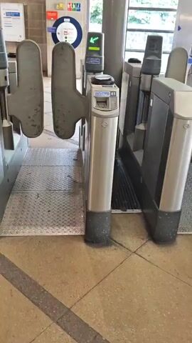 This video shows a close-up view of ticket barriers at what appears to be a train station in Greater London, UK. The barriers are made of metal and plastic, with a patterned metal ramp leading up to one of the gates. The gates themselves are designed to swing open.

A close-up shot focuses on a ticket reader with a contactless payment device labeled "CHASE OYSTER" and instructions to "Please touch your card that you want on the reader". The device has a small orange light on. Another ticket reader is visible to the left, with a green arrow illuminated, indicating it is ready to be used. Text on a blue and white sign above the reader says "Please touch your card that you want on the reader".

In the background, other ticket barriers are visible, as well as people walking through. There are also signs indicating a train station, including a circular sign with a blue and red design, which is characteristic of Transport for London (TfL) signage. The overall impression is of a busy public transport hub. The lighting suggests it is daytime.