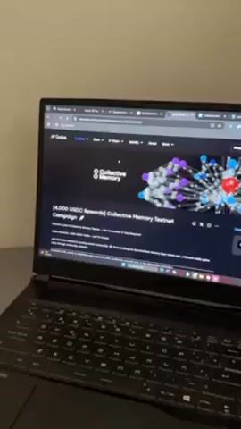 Here is a detailed description of the scene:

A laptop rests on a flat, metallic gray surface, likely a desk or table. The screen displays a webpage with a black background, titled "Collective Memory." Text and images related to a "[4,000 USDC Rewards] Collective Memory Testnet Campaign" are visible. Menu options like "Explore," "Earn," "G Token," "Identity," and "About" are present at the top of the page. The laptop's keyboard is black, and the room's lighting is neutral, casting a slight sheen on the laptop's surface.