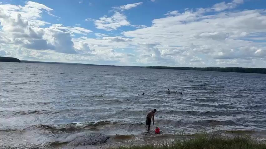 A wide shot shows a lake under a cloudy sky with a few people swimming in the distance. In the foreground, a man wearing swim trunks is wading in the shallows of the lake. He is bent over, appearing to interact with a child in an orange float. The water is choppy and there are waves lapping at the shore. The shoreline is covered in grass and some sand. The background shows a treeline across the lake.