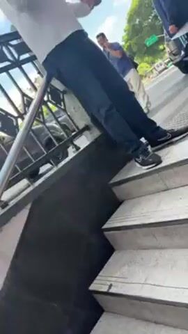 The video begins with a view from stairs leading to a sidewalk, where a person wearing jeans and black athletic shoes stands. There are other people walking on the sidewalk. The camera pans to show people crossing the street at a crosswalk. Cars pass by. The camera then focuses on two people dressed in matching blue shirts with colorful trim. Finally, the camera pans to show a street corner with a small green kiosk advertising coffee and a KFC cafe. Barbed wire is visible above the kiosk. The sky is blue with white clouds.