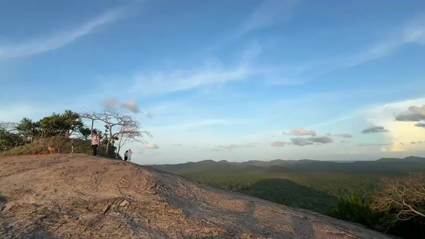 The video shows a scenic view from the top of a rocky hill. The sky is blue with scattered white clouds. In the distance, a vast green forest stretches out to meet the horizon where hills are visible. There are trees and rocks scattered on the hilltop. Initially, two people are visible on the hilltop, one taking a picture of the other. The camera pans to the right revealing a large boulder on the hilltop. Later, a woman in a dress walks into frame on the right.