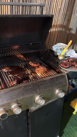 A black gas grill with its lid open sits in front of a bamboo fence. On the grill, eggplants and two patties are cooking. A silver platter containing cooked meat rests on a small table to the right of the grill. A plastic bag and some utensils are also on the table. The grill has three control knobs. The area around the grill is dimly lit.