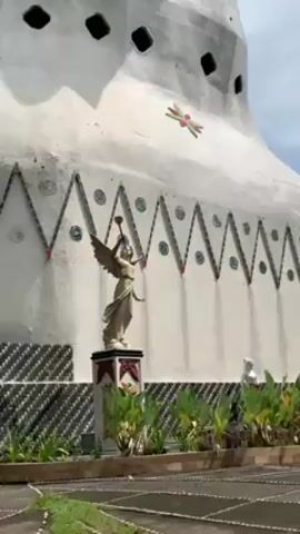 Here is a concise description of the video:

The video showcases a tall, white, chicken-shaped building with a red beak and gold crown. The building is decorated with geometric patterns and small windows. In front of the building, there is a statue of an angel holding a trumpet. Lush greenery surrounds the base of the building and the statue. The camera pans upwards to reveal the full structure of the chicken building against a slightly cloudy sky.