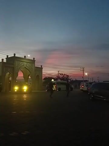 A sunset scene with a sky transitioning from a deep blue to hues of pink and orange. In the foreground, a dark asphalt surface is visible with several cars parked. A large, ornately decorated archway, possibly part of a building or entrance, is illuminated by car headlights. A white van is parked behind the archway, and other vehicles are scattered throughout the scene. Two individuals, silhouetted against the fading light, are walking in the middle ground. The overall atmosphere is serene and tranquil, capturing the beauty of twilight.