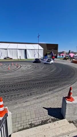 The video shows two cars drifting on a paved surface under a clear blue sky. The cars are leaving tire marks on the ground, indicating a drift event or competition. In the background, there is a large white tent, some spectators, and orange traffic cones marking a course. One car appears to be white with racing decals, and the other is dark-colored. The drifts are creating smoke, suggesting high-performance driving. The scene is an outdoor, possibly urban or event space, environment.