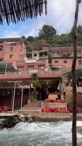 The video shows a man standing on a balcony of a building, dancing. The building is located in a mountainous area with other buildings nearby. There is a stream of water flowing in the foreground. People are visible walking and sitting around the building. The sky is cloudy.