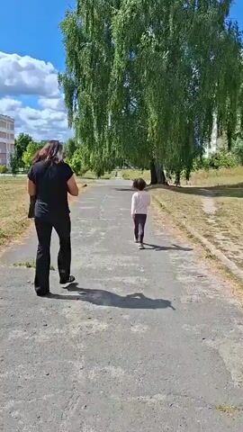 A person in a black outfit and a child in a light-colored top and dark pants walk away from the camera on a paved path. A large tree with drooping branches stands to the right of the path. In the background, a building and more trees are visible under a blue sky with white clouds. The scene appears to be a park or outdoor public space on a sunny day.
