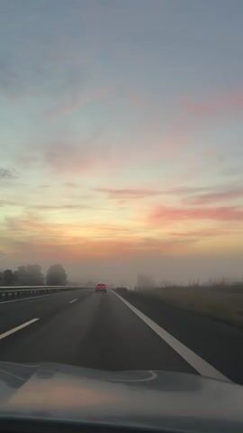 A car is driving down a highway. The sun is setting or rising in the background, casting a pink and orange glow across the sky. There is fog on the highway and in the surrounding fields, obscuring some of the trees and landscape. The interior of another car is visible in the foreground, suggesting the video is being filmed from inside a moving vehicle.