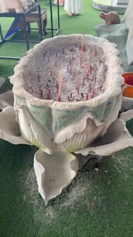 The video shows an incense burner shaped like a lotus flower. The burner is filled with ash and numerous incense sticks, some of which are lit, producing smoke. The burner is placed on a green artificial turf surface. In the background, there are blurred figures of people, possibly in traditional clothing, and other decorative items, including turtle figurines and a white object resembling a shell or a statue of a turtle. The lighting suggests an outdoor or semi-outdoor setting with natural light. The camera is positioned above and looking down at the incense burner.