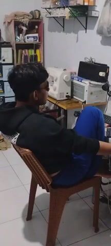 A young person with dark hair, wearing glasses, a black hooded sweatshirt, and blue pants sits in a brown wicker chair, facing a computer monitor. The person's left arm rests on the back of the chair, and their right arm is extended forward, likely towards the keyboard. The background shows a shelf unit filled with various items, including bottles and containers, a white sewing machine, and a white and grey appliance. The room appears to be an indoor space with a tiled floor and a light-colored wall. The lighting is dim.